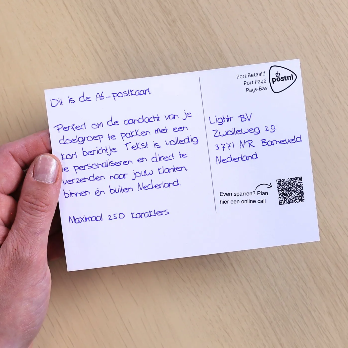 Hand holding an A6 postcard above a wooden table, written with a real pen